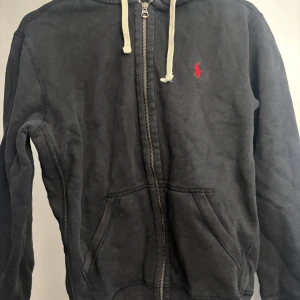 Svart hoodie från Ralph Lauren - Svart hoodie från Ralph Lauren med dragkedja, vit snörning i huvan och röd broderad logga på bröstet. Mjuk bomullskänsla och kängurufickor framtill. Perfekt för chill dagar eller när du vill ha en clean streetstyle.