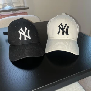 Två kepsar svart och begie  - Säljer två klassiska kepsar stadium, en i svart med vit broderad logga och en i begie med svart broderad logga. Båda har böjd skärm och justerbar rem baktill. Perfekt accessoar för en sportig och trendig look. Båda för 600kr.