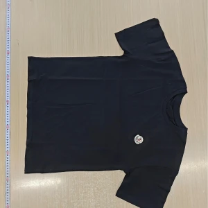 Svart t-shirt från Moncler - Svart t-shirt från Moncler med klassisk rund hals och korta ärmar. På bröstet sitter Monclers ikoniska logotyp-patch. T-shirten är gjord i mjuk bomull och har en clean, stilren look som passar perfekt till jeans eller shorts.