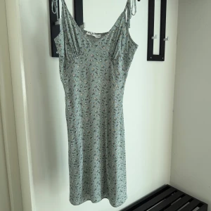 Blommig ljusgrön klänning från NA-KD - Supersöt ljusgrön klänning från NA-KD med små blommor. Modellen är kort med smala axelband som kan knytas och har smock i ryggen för extra komfort. 