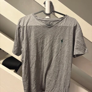 Grå v-ringad t-shirt Polo Ralph Lauren - Grå v-ringad t-shirt från Polo Ralph Lauren i mjuk bomull. Klassisk kortärmad modell med broderad grön logga på bröstet. Perfekt basic med clean look och skön passform. Snygg att matcha med jeans eller shorts.