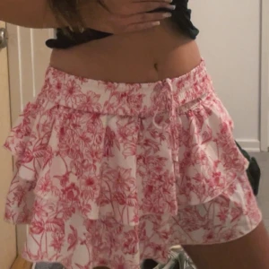 Rosa blommig volangkjol - Supersöt kort kjol med rosa blommigt mönster på vit botten. Kjolen har flera volanger och bred resår i midjan för en bekväm passform.