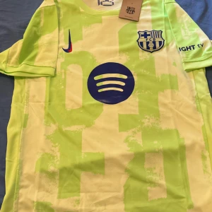 Barcelona kortärmad fotbollströja Nike - Säljer en Barcelona fotbollströja från Nike med coolt ljusgult och ljusgrönt mönster. Tröjan har korta ärmar, FCB-logga på bröstet, Nike Swoosh i rött och blått samt Spotify-logga i mörkblått. Perfekt för dig som vill sticka ut på planen eller läktaren.
