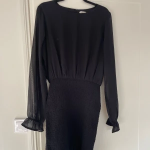 Svart kort klänning med volang - Snygg svart klänning med långärmade transparenta ärmar och volangdetaljer vid ärmslut och nederkant. Smockad midja och kjol ger en cool och figurnära siluett. Perfekt för dig som gillar stilrena och trendiga plagg med lite extra detaljer. Storlek 170 funkar bra som xs/s