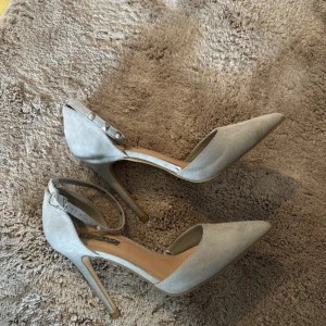Ljusgrå pumps med spetsig tå och nitar - Snygga ljusgrå pumps med spetsig tå och hög klack. Skorna har en smal ankelrem med coola silverfärgade nitar och pärldetaljer. Tillverkade i mockaliknande material som ger en lyxig känsla. Perfekta för dig som vill sticka ut med en stilren men edgy look.