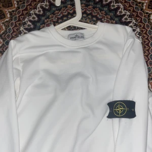 Vit långärmad Stone Island tröja - Vit långärmad tröja från Stone Island med klassisk rund hals och ikonisk patch på vänster ärm. Tröjan är i mjuk bomull och har en clean, stilren look som passar perfekt till streetwear eller casual outfits.