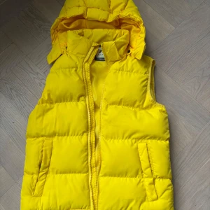 Gul dunväst från McKinley med huva - Snygg gul dunväst från McKinley med avtagbar huva och dragkedja framtill. Västen har två fickor på sidorna och är quiltad för extra värme. Perfekt för lager-på-lager och en färgklick till din outfit.
