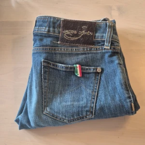 Jacob cohen jeans  - Snygga blå jeansbyxor från Jacob Cohen med klassisk femficksdesign och en liten italiensk flaggdetalj på bakfickan. Byxorna har rak passform och normal midja, perfekta till vardagslooken. 