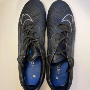 Nike Phantom FG/AG  - Säljer ett par Nike Phantom fotbollsskor i svart med blå detaljer och silvriga swoosh-loggor. Skorna har snörning, mönstrad ovandel och platt sula med dobbar för bra grepp på planen. Perfekta för dig som vill sticka ut på matchen!