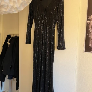 Svart glittrig långklänning  - Svart långklänning med glittriga paljetter från In The Style. Klänningen har v-ringning med snörning framtill, långa ärmar och figurnära passform. strl M ej anvönd