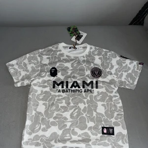 BAPE x Inter Miami camo t-shirt - Snygg t-shirt från BAPE i samarbete med Inter Miami. T-shirten har grått och vitt camouflagemönster, tryck med MIAMI och BAPE 93, samt broderade patches. Klassisk passform och tillverkad i mjuk bomull. Perfekt för dig som gillar streetwear och fotboll.
