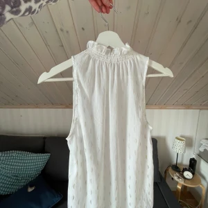 Vit ärmlös blus från Mango - Supersöt vit ärmlös blus från Mango med guldiga detaljer i tyget. Blusen har hög krage med volang och smock, samt knäppning framtill. Tunt och luftigt material som känns lyxigt och passar perfekt till varmare dagar.