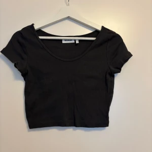 Svart croppad topp från Weekday - En svart, croppad topp från Weekday med korta ärmar och rund halsringning. Toppen är ribbad och har en tight passform som sitter snyggt på kroppen. Perfekt att styla med höga jeans eller kjol för en clean look.