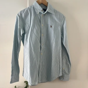 Randig skjorta från Ralph Lauren - Klassisk skjorta från Ralph Lauren i vitt och blått med tunna ränder. Custom fit-modell med lång ärm, knappar framtill och broderad logga på bröstet. Perfekt för en stilren och tidlös look.