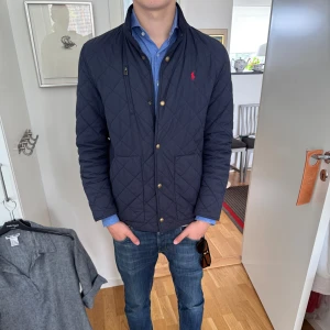  Polo Ralph Lauren jacka  - Snygg mörkblå tunn jacka från Polo Ralph Lauren med klassisk passform. Jackan har guldiga knappar, dragkedja och diskret röd logga på bröstet. Perfekt för dig som vill ha en stilren och tidlös look. Fodrad och med två fickor framtill. 