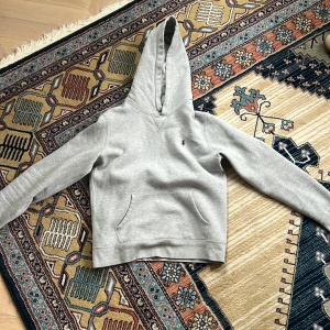 Grå hoodie från Polo Ralph Lauren - Snygg grå hoodie från Polo Ralph Lauren med klassisk huva och magficka. Tröjan har en liten broderad logga på bröstet och är tillverkad i mjuk bomullsblandning. Perfekt för en chill och stilren look.