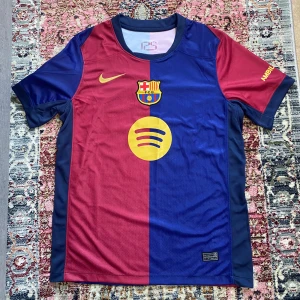 FC Barcelona Lamine Yamal tröja Nike S - FC Barcelona matchtröja från Nike med Lamine Yamal och nummer 19 på ryggen. Halva tröjan är vinröd och andra halvan marinblå, med gula detaljer och klubbmärke på bröstet. Tillverkad i Dri-FIT-material för bästa komfort. Klassisk rund hals och korta ärmar.