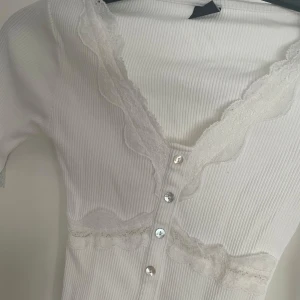 Vit ribbad blus med spets och knappar - Supersöt vit ribbad blus med v-ringning, spetsdetaljer längs halsringning och midja samt silvriga knappar framtill. Långärmad och figurnära passform, i nyskick! 