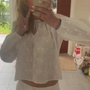 Vit blommig blus med knytning - Supersöt vit blus med broderade blommor och knytning vid halsen. Lätt transparent tyg, långa ärmar med resår vid handlederna och avslappnad passform. Perfekt för en fräsch och somrig look!