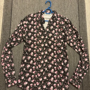 Blommig skjorta från The Blue Uniform - Svart skjorta med rosa och vita blommor från The Blue Uniform. Långärmad modell med knappar framtill och klassisk krage. Tyg i mjuk bomull och liten broderad ankare på bröstet. Perfekt för dig som gillar unika mönster och vill sticka ut.