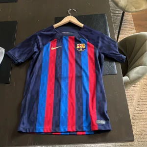 Barça matchtröja Nike blå/röd - FC Barcelona matchtröja från Nike med klassiska blå och röda vertikala ränder. Tröjan har klubbmärke på bröstet och Nike-logga i guld. Tillverkad i lätt och ventilerande material, perfekt för fotboll eller att bära som supporter.