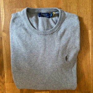 Grå tröja från Ralph lauren  - Klassisk grå tröja från Polo Ralph Lauren med rund halsringning och diskret broderad logga på bröstet. Tröjan är stickad i mjuk bomull och har långa ärmar samt ribbade muddar vid ärmslut och nederkant. Perfekt för lager-på-lager-stil.