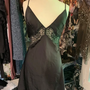 Svart spetsklänning från Hunkemöller - Supersnygg svart kort klänning från Hunkemöller med tunna axelband, djup v-ringning och spetsdetaljer över midjan. Klänningen har öppen rygg med snörning och är tillverkad i ett glansigt, mjukt material. Skulle tro att det är storlek M men även s eftersom man kan justera snörena i ryggen.