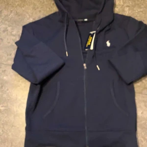 Polo Ralph Lauren zip - Snygg marinblå hoodie från Polo Ralph Lauren med dragkedja och huva. Klassisk logga broderad på bröstet och snörning i huvan. Tillverkad i mjuk bomull, perfekt för chill dagar. Fickor framtill och detaljer i silver på dragkedjan.