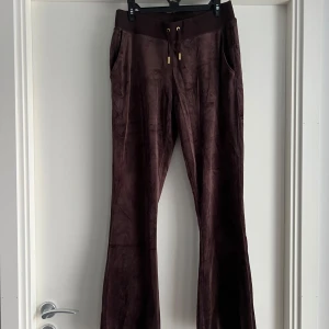 Bruna velourbyxor Juicy Couture M - Mörkbruna mjukisbyxor från Juicy Couture i velour med bootcut-snitt. Byxorna har snörning i midjan med guldfärgade detaljer och fickor på sidorna. Perfekta för en chill och trendig look. 