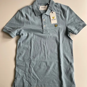 Ljusblå piké från Lyle & Scott - Snygg ljusblå pikétröja från Lyle & Scott med klassisk krage, korta ärmar och knäppning framtill. Tröjan har den ikoniska gula loggan broderad på bröstet och är gjord i mjuk bomull. Perfekt för en clean och stilren look. Aldrig använd och tag sitter kvar, säljer för 200kr