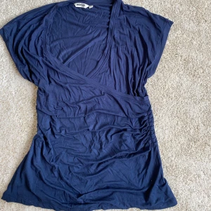 Mörkblå omlottopp från Indiska - Mörkblå omlottopp från Indiska i mjuk bomull. Toppen har v-ringning, korta ärmar och rynkade detaljer i sidan för en snygg passform. Perfekt att styla med jeans eller kjol för en chill look.