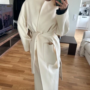 Beige kappa med bälte och fickor - Stilren beige kappa i lång modell med raka ärmar och stora fickor framtill. Knyts i midjan med bälte för en snygg siluett. Perfekt för dig som gillar minimalistisk och clean look. Rund halsringning och slits i sidan.