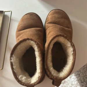 Bruna UGG boots i mocka med fårskinn - Mysiga bruna UGG boots i klassisk modell med mjukt fårskinnsfoder och ovandel i mocka. Skorna har platt sula och rund tå, perfekta för kalla dagar. UGG-logga baktill och kort skaft för en chill look.