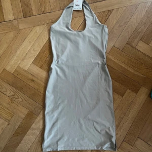 Beige halterneckklänning SOFT 2000 - Snygg beige halterneckklänning från SOFT 2000 MILLENNIUM. Klänningen är kort, figurnära och har en djup urringning framtill. Tillverkad i mjukt, stretchigt material som känns skönt mot huden. Perfekt för dig som gillar minimalistisk och stilren look.