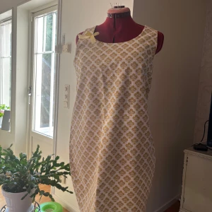 Gulblommig ärmlös kortklänning med rosett - Supersöt ärmlös kortklänning med gult blommönster på vit botten och en söt gul rosett vid axeln. Klänningen har en rak passform och är perfekt för varma dagar. Materialet känns lätt och luftigt, vilket gör den skön att bära.