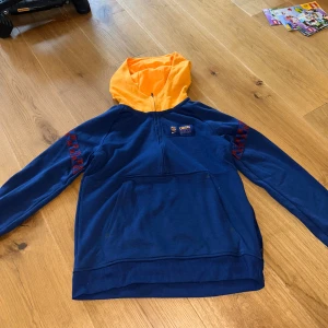 Nike blå hoodie med orange huva - Nike barcelona hoodie i blått med orange huva och halv dragkedja. Tröjan har röda rutiga detaljer på ärmarna, stor känguruficka och är tillverkad i Dri-FIT polyester. Loose fit för en avslappnad stil. Perfekt för dig som gillar sportig vibe.