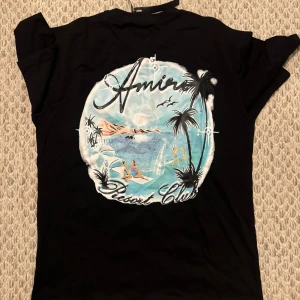 Oversize Amiri tshirt - Svart t-shirt från Amiri med stor färgglad print på ryggen som visar en strand, palmer och surfare. Snyggt tryck med texten 'Amiri Resort Club' i vitt och blått. Klassisk passform och korta ärmar, perfekt för en chill look.