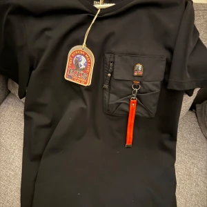 Svart t-shirt från Parajumpers med ficka äkta - Svart t-shirt från Parajumpers med cool bröstficka som har dragkedja, knapp och en orange detalj. T-shirten har korta ärmar och en snygg logga på fickan. Perfekt för dig som gillar streetwear och vill sticka ut lite extra.
