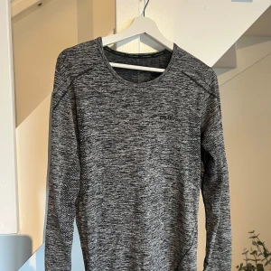 Grå långärmad träningströja/underställ Craft  - Superskön långärmad träningströja från Craft i gråmelerat mönster. Tröjan har rund halsringning, smidig passform och är tillverkad i ett lätt och stretchigt syntetmaterial som andas. Perfekt för löpning eller gymmet. Funkar även perfekt som underställ på vintern. 
