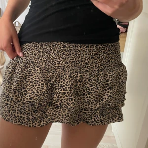 Leopardmönstrad kortkjol med volang - Supersöt kortkjol med leopardmönster och volangkant. Kjolen har smock i midjan för en bekväm och flexibel passform. Färgerna går i beige och svart, vilket gör den lätt att matcha med både enkla och mer statement toppar. Perfekt för dig som gillar trendiga och lekfulla plagg.