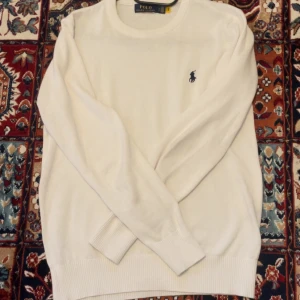 Vit stickad tröja från Polo Ralph Lauren - Vit stickad tröja från Polo Ralph Lauren med klassisk rund hals och broderad marinblå logga på bröstet. Tröjan har långa ärmar och ribbade muddar vid ärmslut och nederkant. Perfekt för en clean och stilren look.