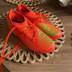 Röda Adidas fotbollsskor med neonmönster - Snygga fotbollsskor från Adidas i röd färg med neongröna detaljer och mönster. Skorna har snörning och dobbar i grönt och rött för bra grepp på planen. Lätta och smidiga med en sportig look som sticker ut. Och dom har bara används en gång.