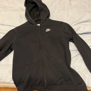Svart Nike hoodie med dragkedja XL barn - Svart hoodie från Nike i storlek XL med vit logga på bröstet. Tröjan har huva, hel dragkedja och två stora fickor, passar xs/s