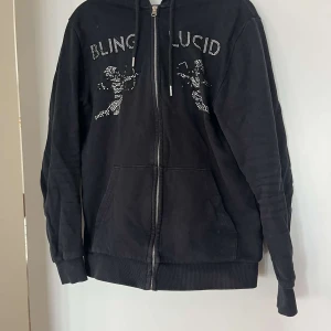 Svart hoodie med strass från Blinglucid - Svart hoodie från Blinglucid i storlek XS med dragkedja, huva och snörning. Framsidan har coola strassdetaljer med texten BLINGLUCID och två figurer. Perfekt för dig som gillar streetstyle och vill sticka ut.