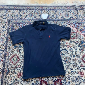 Mörkblå pikétröja från Ralph Lauren - Snygg mörkblå pikétröja från Ralph Lauren med klassisk krage och två knappar. Tröjan har den ikoniska röda polospelaren broderad på bröstet och är i bomull med en slim fit-passform. Perfekt för en clean och stilren look.