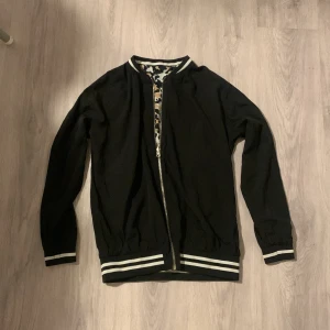 Leopardmönstrad bomberjacka med dragkedja - Snygg bomberjacka med leopardmönster i svart, beige och brunt. Jackan har svarta muddar med vita ränder vid ärmslut och midja samt hel dragkedja framtill. Insidan är svart, vilket gör den vändbar för två olika looks. Perfekt för dig som vill sticka ut.