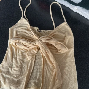 Beige linnetopp med stor rosett från Gina tricot - Supersöt beige linnetopp med smala axelband och en stor knytrosett framtill. Toppen är ärmlös och har en lätt transparent känsla i ett mjukt, tunt material. Perfekt för varma dagar när du vill ha en chill och trendig look.