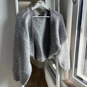 Grå stickad bolero från Gina Tricot - Mysig grå stickad bolero från Gina Tricot med ribbad struktur och avslappnad passform. Koftan är öppen framtill och har långa ärmar, perfekt att slänga på sig över en t-shirt eller linne. Enkel och stilren design som funkar till det mesta. 