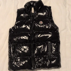 De en snygg Moncler vest har för många kläder vill inte ha den längre - Svart, glansig dunväst från Moncler med hög krage och dragkedja framtill. Västen har två fickor med dragkedja och ikonisk Moncler-logga på bröstet. Perfekt för lager-på-lager och ger en clean, sportig vibe. Den är i perfekt condition 