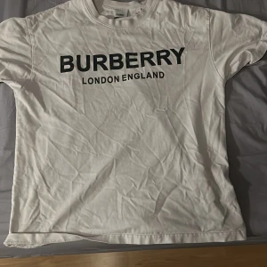 Vit Burberry t-shirt med tryck - Behöver bara strykas! Vit t-shirt från Burberry med klassiskt svart logotryck 'BURBERRY LONDON ENGLAND' på bröstet. T-shirten har rund hals, korta ärmar och är gjord i mjuk bomull. Enkel och stilren design som passar perfekt till jeans eller shorts.